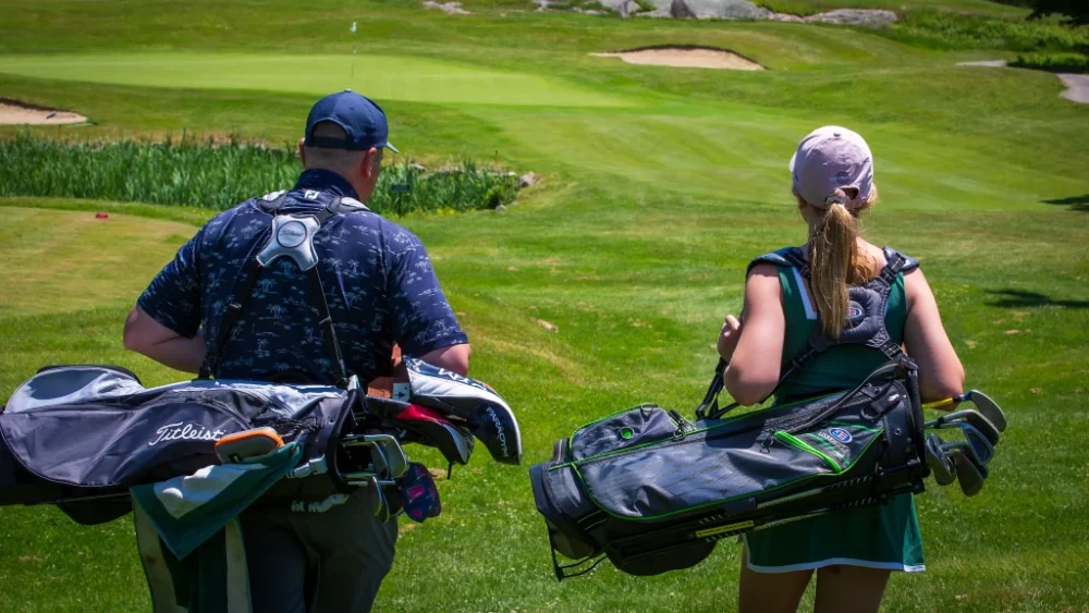 a young woman and an man on the golf course with their caddies walking to a hole